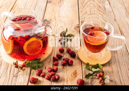 una grande teiera di vetro con una decozione di bacche di rosa e tè in una tazza di vetro con un doppio fondo su un tavolo di legno tra bacche secche Foto Stock