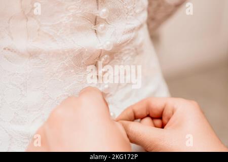 Mattina preparazione della sposa. Primo piano di una bridesmaid, allacciando un sacco di bottoni sul vestito di nozze della sposa. La sposa in un matrimonio bianco Foto Stock