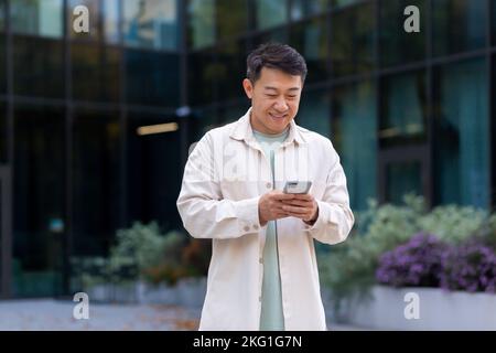Uomo asiatico che utilizza il telefono fuori ufficio edificio, libero uomo d'affari digitando messaggio e sorridendo, programmatore in abiti casual navigazione pagine web online da app. Foto Stock