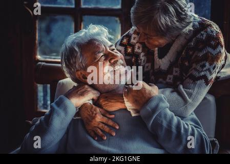 Ritratto di coppia di anziani innamorati a casa nella luce oscura della sera. L'uomo anziano sorride alla moglie di sua donna matura abbracciando e godendo relationshi Foto Stock