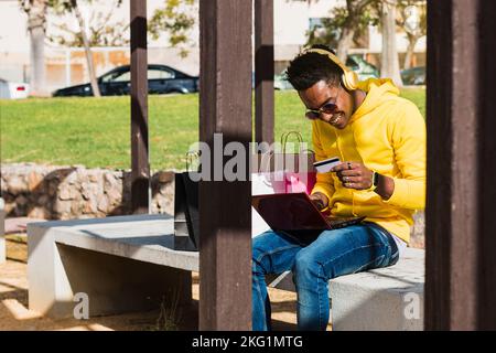 Un giovane africano sorridente siede su una panca del parco vicino ad alcune borse della spesa usando un computer portatile e tenendo una scheda di pagamento mentre indossa lui giallo Foto Stock