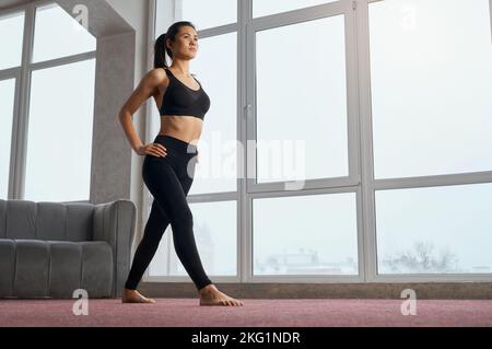 Vista laterale di una donna sportiva e sottile in piedi con fianchi a mani libere in interni. Giovane bruna donna che fa un passo avanti a piedi nudi, praticando yoga. Concetto di armonia ed equilibrio interiore. Foto Stock