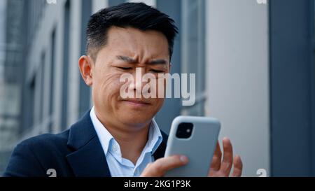 Primo piano arrabbiato preoccupato sottolineato asiatico di mezza età 40s uomo utente imprenditore datore di lavoro imprenditore uomo cercando di navigare sul telefono cellulare rotto all'aperto in città Foto Stock