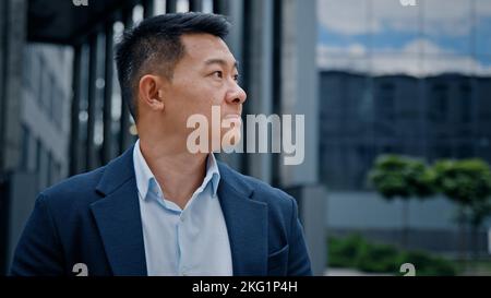 Pensivo datore di lavoro premuroso guardare lontano pensare sognare girare a fotocamera sorridere in città all'aperto uomo asiatico coreano capo d'affari leader società CEO Foto Stock