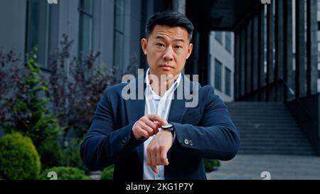 Arrabbiato preoccupato 40s asiatico uomo d'affari maschio frustrato boss manager CEO in attesa di riunione all'aperto in città nervosa fastidio frustrato indicando sopra Foto Stock