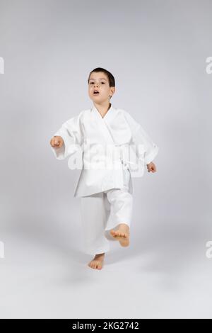 Un ragazzino in un kimono pratica il karate su uno sfondo bianco, calciando in avanti. Foto Stock