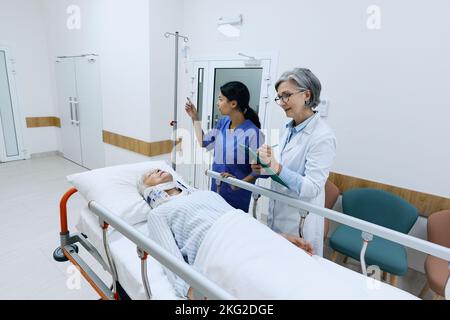 medico esaminando paziente anziano con lesioni al collo disteso su una barella medica con collare cervicale in corridoio ospedaliero, preso in ambulanza Foto Stock