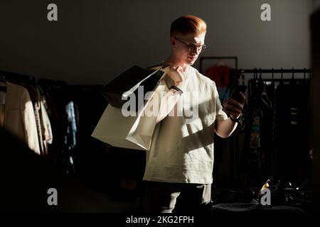 Moody ritratto di gen Z giovane uomo utilizzando lo smartphone mentre si gode lo shopping al negozio di Thrift in luce del sole e tenendo sacchetti di carta, spazio copia Foto Stock