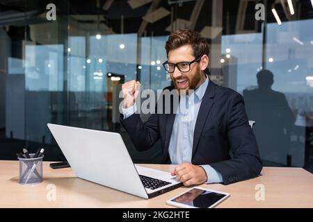 Uomo d'affari di successo che celebra la vittoria e il buon successo aziendale, il capo che utilizza il notebook per leggere la notifica dallo schermo e tenere la mano su un gesto trionfale sorridente. Foto Stock