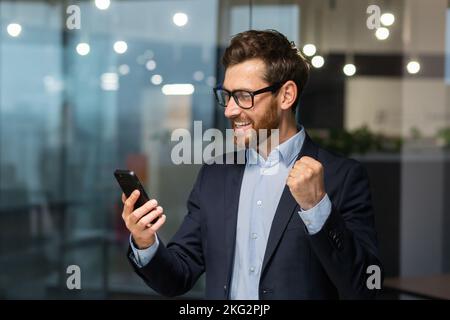 Investitore riuscito dell'uomo d'affari usando il telefono, l'uomo che festeggia la vittoria ed il successo di successo, tenendo la mano in su il gesto di trionfo, il boss nel vestito di affari all'interno dell'ufficio in piedi vicino alla finestra. Foto Stock