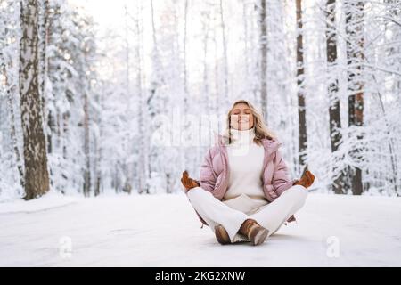 Giovane donna con capelli biondi in abiti invernali che si diverte a sedere nella foresta invernale innevata Foto Stock