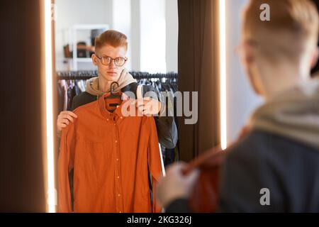 Ritratto di Moody di giovane uomo che prova su vestiti in camerino al negozio di Thrift e guardando lo specchio Foto Stock