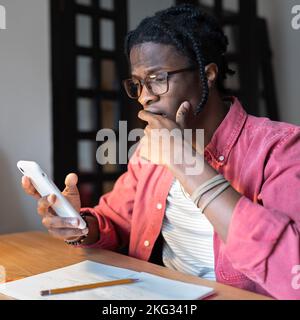 Frustrato uomo afro-americano spaventato siede a tavola con il telefono cellulare che copre la bocca con la mano Foto Stock