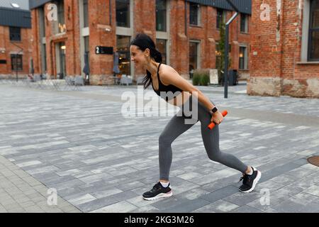 Allegra donna atletica in abbigliamento sportivo che fa affondi con manubri sulla strada della città Foto Stock
