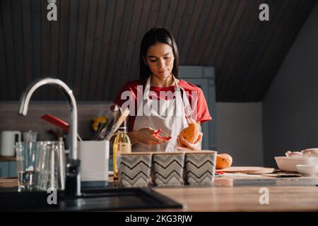 Giovane donna ispanica che sbuccia la patata dolce mentre cucinava in cucina a casa Foto Stock