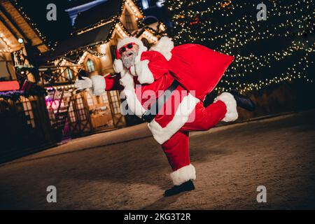 Full size ritratto di overjoyed funky Santa Hold età presenta sacco divertirsi buon umore christmastime decorazione luci all'aperto Foto Stock