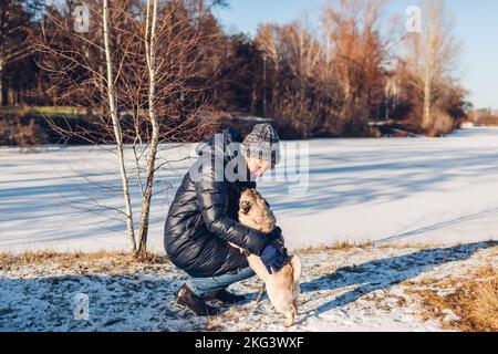 Donna che cammina pug dog in neve parco invernale da ghiaccio lago hugging animale domestico. Imbracatura da cucciolo felice Foto Stock