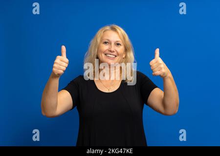 Donna bionda caucasica in abbigliamento casual approvando gesto mano positivo, pollici su sorridente e felice per il successo. Gesto vincente. Isolato su blu Foto Stock