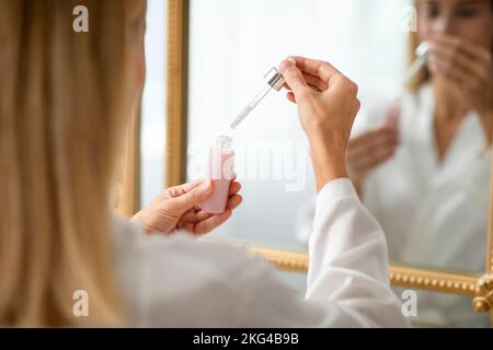 Donna matura irriconoscibile gocciolando siero idratante, pronto per il trattamento di bellezza, in piedi di fronte allo specchio Foto Stock