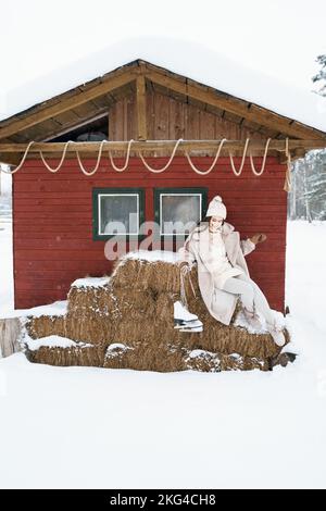 Giovane bella ragazza su balle di fieno vicino fienile con pattini. Inverno freddo innevato in campagna. Camminare, divertirsi, ridere in abiti eleganti, pelliccia co Foto Stock