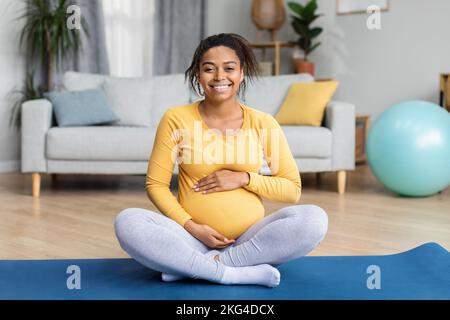 Allegro giovane donna incinta nera con grande pancia pratica yoga sul tappeto nel moderno soggiorno interno Foto Stock