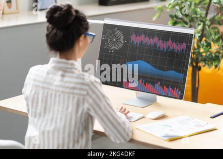 Giovane donna d'affari che analizza le statistiche sullo schermo del computer, lavorando con grafici finanziari e grafici online in ufficio Foto Stock