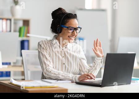 Una lavoratrice entusiasta che effettua videochiamate tramite computer portatile, parla con la webcam e sventura, seduto sul posto di lavoro in ufficio Foto Stock