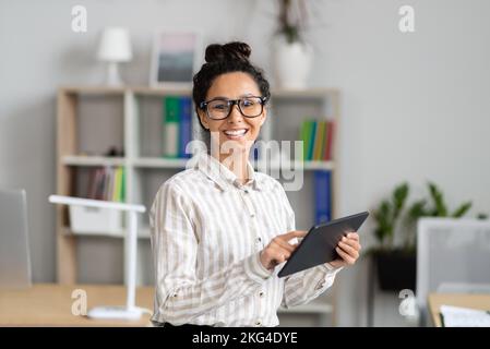 Ritratto di una felice lavoratrice che tiene un tablet digitale, sorride alla fotocamera, in piedi vicino al suo posto di lavoro in ufficio Foto Stock