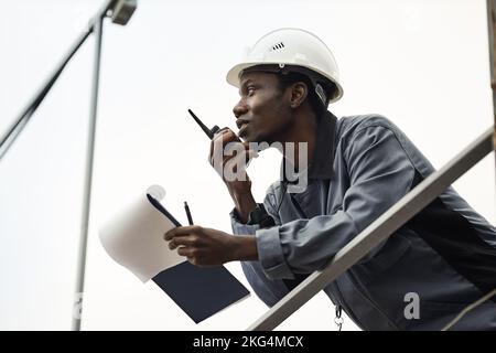 Ritratto grafico dell'operaio maschio nero che indossa l'hardHat e che usa la radio contro il cielo bianco Foto Stock