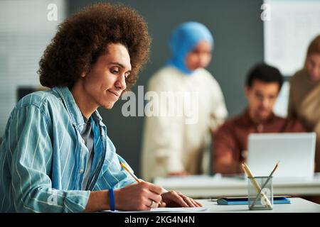 Vista laterale di un giovane studente multietnico che guarda la carta con test grammaticale e la indica mentre pensa alla risposta giusta alla lezione Foto Stock