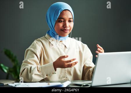 Giovane donna musulmana fiduciosa in casualwear e hijab blu che spiega qualcosa mentre si siede di fronte al notebook durante la lezione online Foto Stock