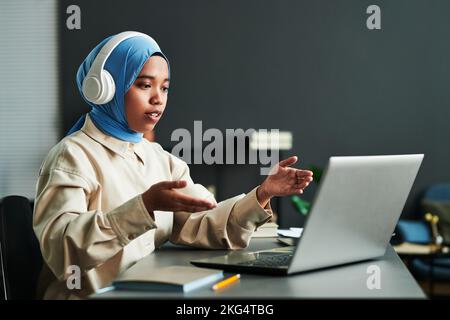 Giovane donna musulmana fiduciosa in cuffie sopra hijab blu che spiega qualcosa durante la comunicazione online Foto Stock