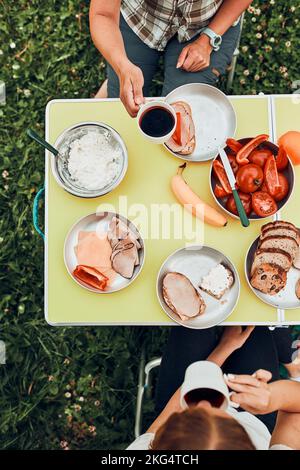 Famiglia che ha la colazione all'aperto in campeggio durante le vacanze estive. Pane, formaggio di ricotta, carne fredda, pomodori, frutta e tazze di caffè sul tavolo. Chiudi Foto Stock