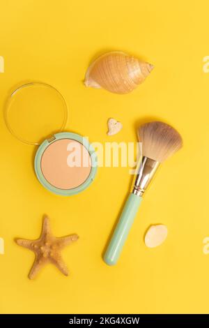 Concetto di trucco estivo. Pennello cosmetico e polvere per il viso con conchiglie e stelle marine su fondo giallo. Foto Stock