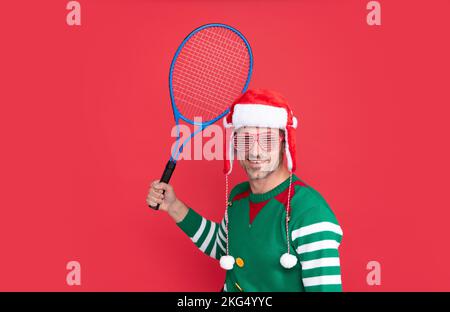 Buon Natale. felice uomo in costume di elfo e cappello di babbo natale. xmas guy tenere il razzo da tennis su sfondo rosso. Felice Anno Nuovo. Foto Stock