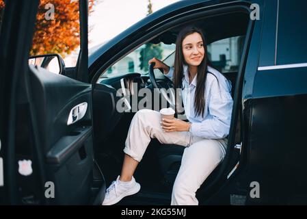 Una donna felice e attraente che tiene una tazza di caffè e che esce dalla sua auto moderna Foto Stock