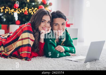 Foto immagine di bello carino simpatico sorridente giovani vogliono scegliere più regali di natale sdraiato piano vicino abete utilizzare computer online consegna al chiuso Foto Stock