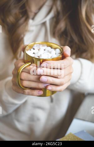 Closeup mani femminili che tengono la tazza dorata di lusso piena di marshmallow cacao Natale atmosfera Foto Stock