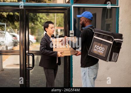 Pizzeria corriere che dà al cliente pizzeria ordine all'aperto, donna che prende pasto pacchetti pila per i colleghi. Uomo afro-americano che consegna il pranzo da asporto, dipendente dell'azienda che riceve fastfood Foto Stock