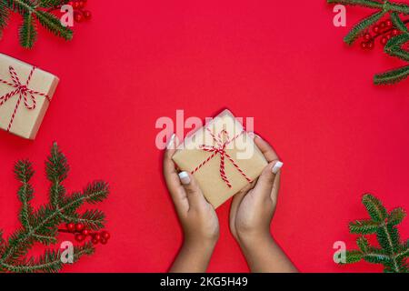 Design creativo sfondo rosso e rami di alberi di Natale, regali, canne caramelle e mani femminili che tengono un regalo di Natale. Foto Stock