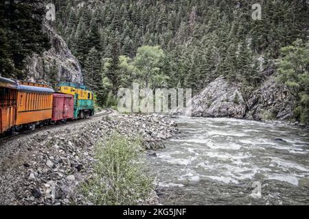 Motore a vapore che circonda una curva con scogliera di montagna su un lato e fiume di corsa sull'altro in montagne boschive di pino - filtro vintage Foto Stock