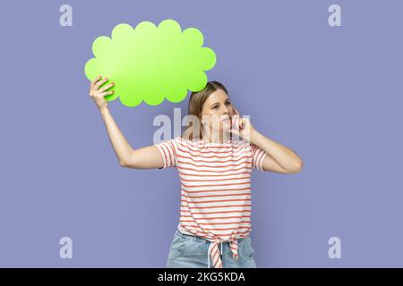 Ritratto di giovane donna bionda pensosa giovane adulto con T-shirt a righe bolla verde per il testo, guardando lontano e tenendo il mento. Studio al coperto isolato su sfondo viola. Foto Stock