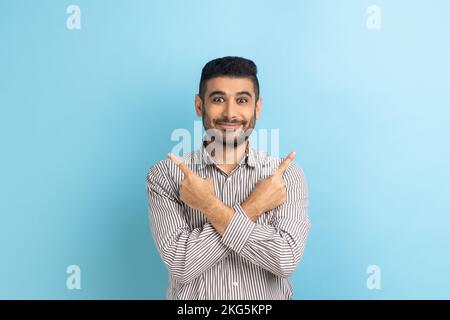 Uomo d'affari sorridente bearded che punta entrambi i lati, mostrando spazio vuoto della copia per la presentazione dell'idea, testo commerciale, sorridente felicemente, portando la camicia a strisce. Studio in interni isolato su sfondo blu Foto Stock