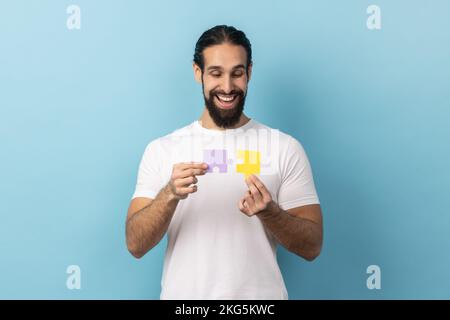 Ritratto dell'uomo con barba in T-shirt che tiene due parti di puzzle e sorridente gioiosamente, pronto a collegare pezzi di puzzle, simbolo di Unione e associazione. Studio in interni isolato su sfondo blu. Foto Stock