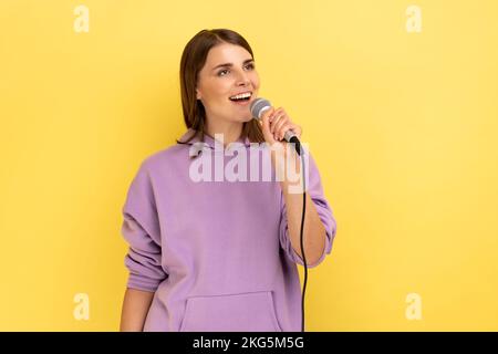 Ritratto di donna positiva cantando forte canzone, tenendo il microfono in mano, divertirsi riposando in karaoke, cantante, indossando felpa con cappuccio viola. Studio al coperto isolato su sfondo giallo. Foto Stock