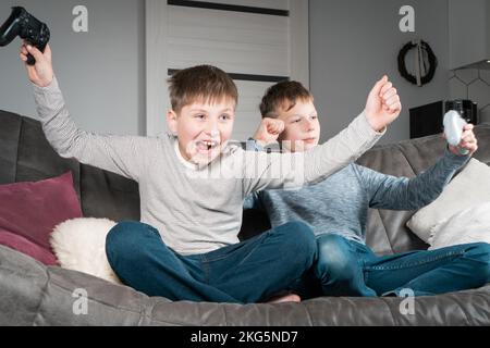 Ritratto di due allegri ragazzi adolescenti seduti sul divano grigio di casa, con joystick del controller di gioco, videogiochi, festeggiamenti Foto Stock