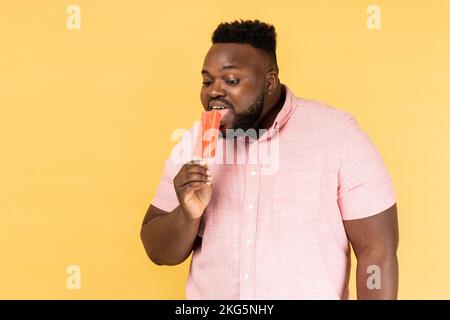 Ritratto di uomo ottimista positivo indossando camicia rosa che tiene leccare gelato dolce, cercando delizioso dessert pasticceria. Studio al coperto isolato su sfondo giallo. Foto Stock