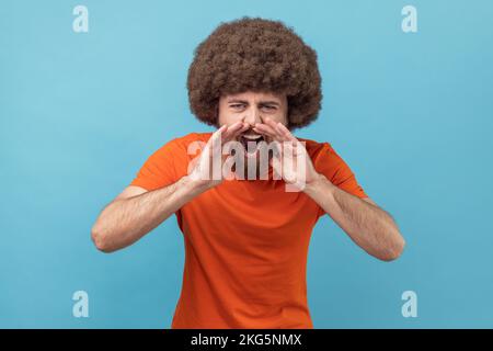 Ritratto di un uomo aggressivo arrabbiato con acconciatura afro che indossa una T-shirt arancione in piedi, tenendo le braccia vicino alla bocca aperta e urlando. Studio in interni isolato su sfondo blu. Foto Stock