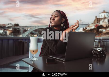 Ritratto di una giovane donna d'affari africana abbagliante con un computer portatile e un cocktail in un caffè di strada; ravishing imprenditore nero donna con trecce rosse i Foto Stock