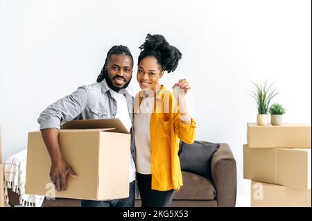 Moderno eccitato marito e moglie, in piedi nel loro nuovo salotto, l'uomo che tiene la scatola grande, donna che tiene le chiavi, guardando la macchina fotografica, sorridendo. Felice coppia afro-americana si trasferisce nella loro nuova casa Foto Stock